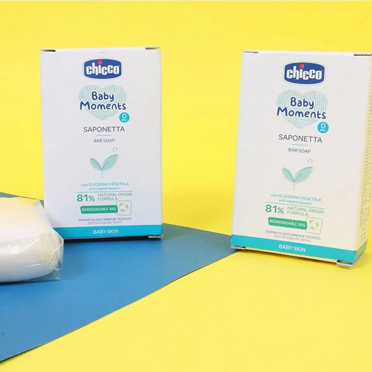 دو جعبه‌ی صابون Baby Moments چیکو و یک قالب صابون در کیسه‌ی شفاف روی زمینه‌ی زرد و آبی