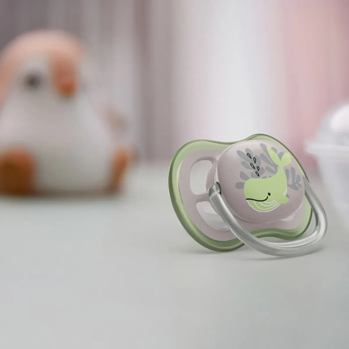 پستانک فیلیپس اونت Ultra Air با طرح نهنگ سبز روی سطح خاکستری، با عروسک محو در پس‌زمینه