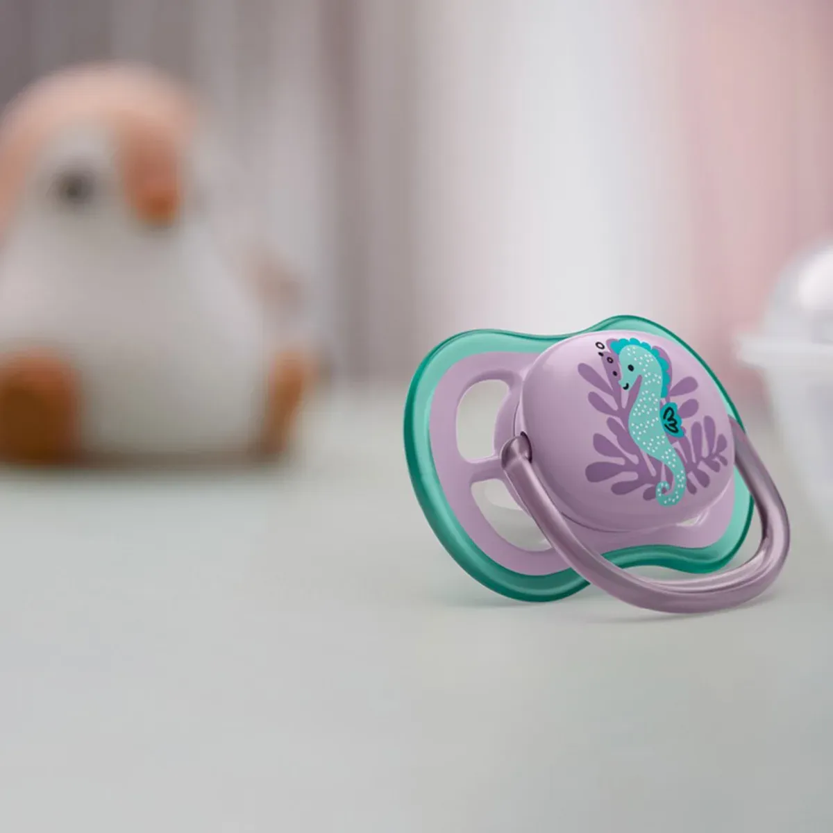 پستانک فیلیپس اونت Ultra Air با طرح اسب دریایی فیروزه‌ای روی زمینه‌ی بنفش، با عروسک محو در پس‌زمینه
