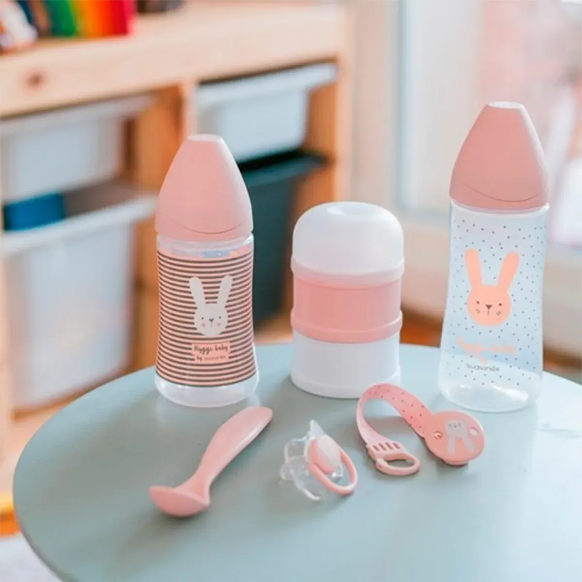 دو شیشه شیر صورتی طرح خرگوش، محفظه‌ی شیرخشک، قاشق، پستانک و بند پستانک ست Welcome Baby روی میز